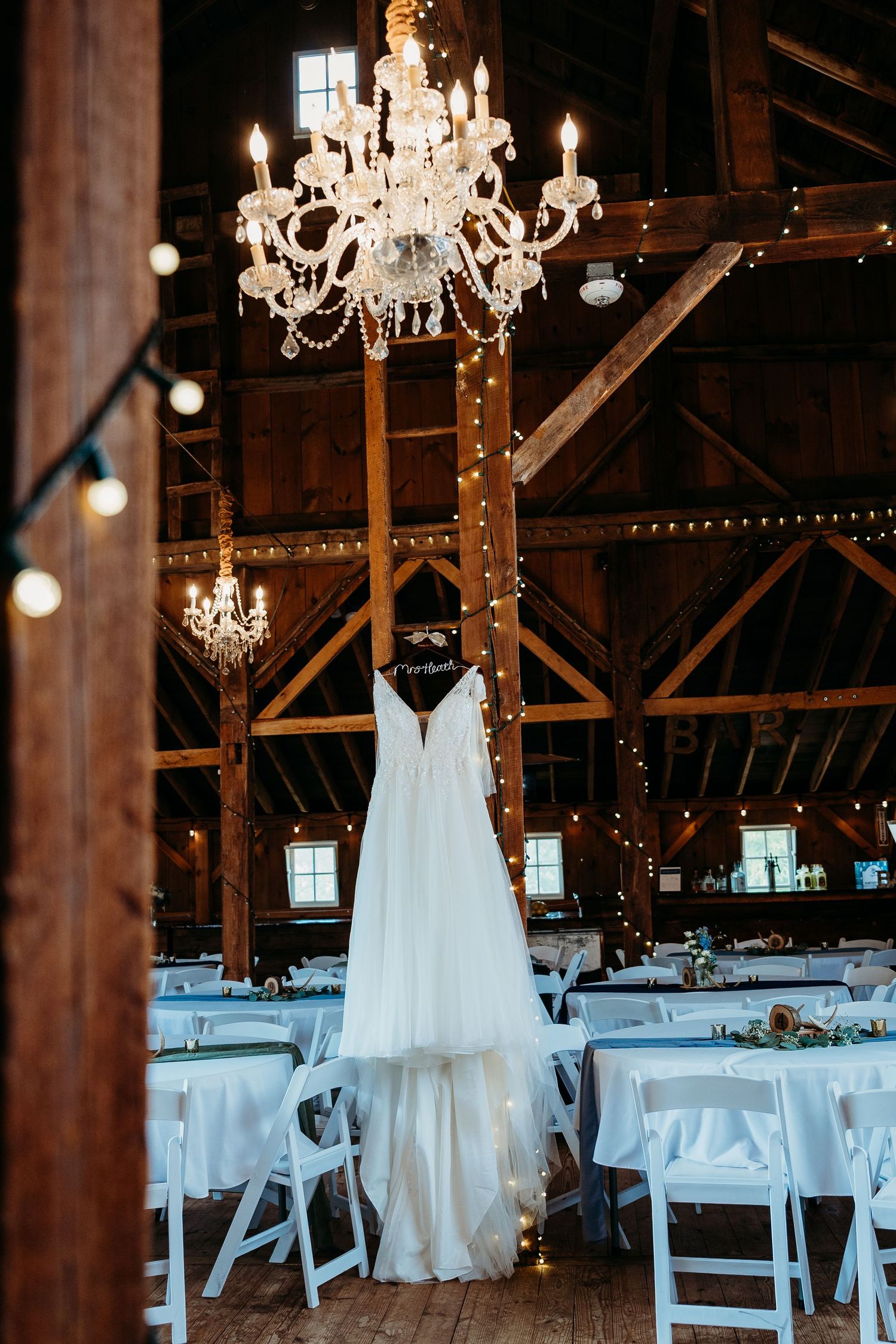 Wedding dress hangs elegantly in rustic barn venue with crystal chandeliers and white table settings.