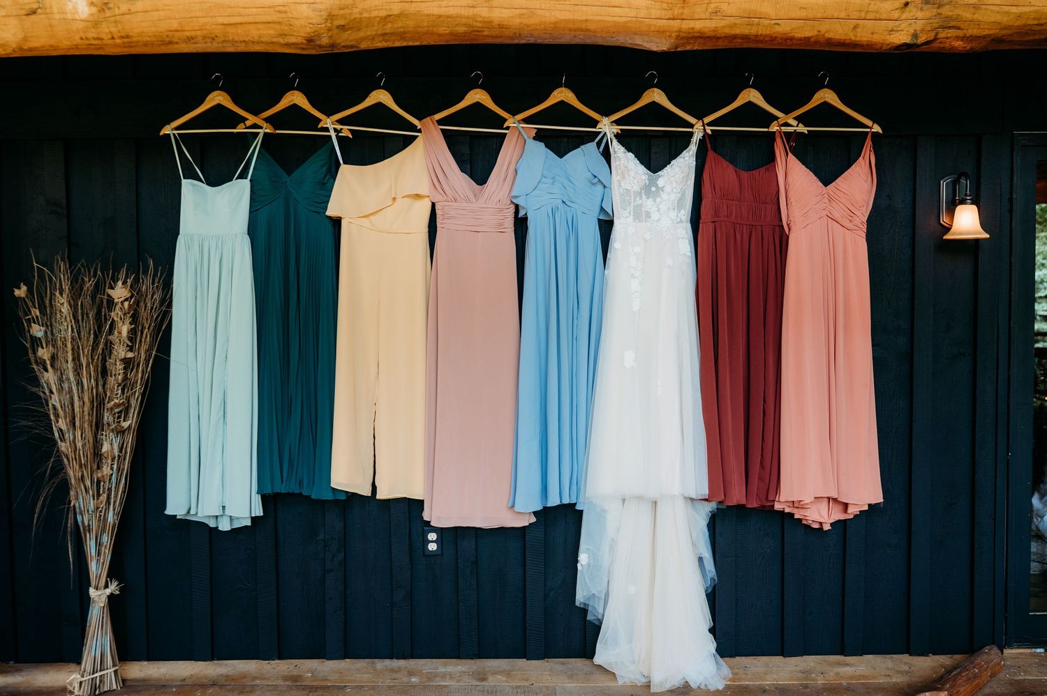 Wedding dresses in soft pastel colors hang in a row against a dark navy wall with dried flowers on display.