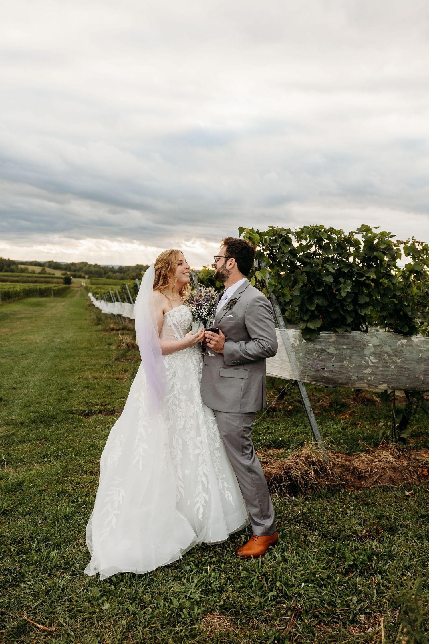 A couple shares a romantic moment in their wedding attire while standing in a scenic vineyard at sunset.