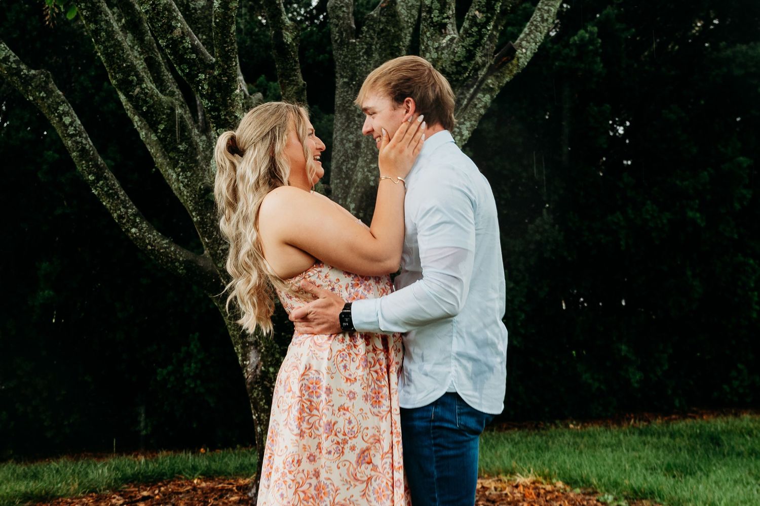 Couple embracing and smiling at each other outdoors near a large moss-covered tree in a lush green garden.