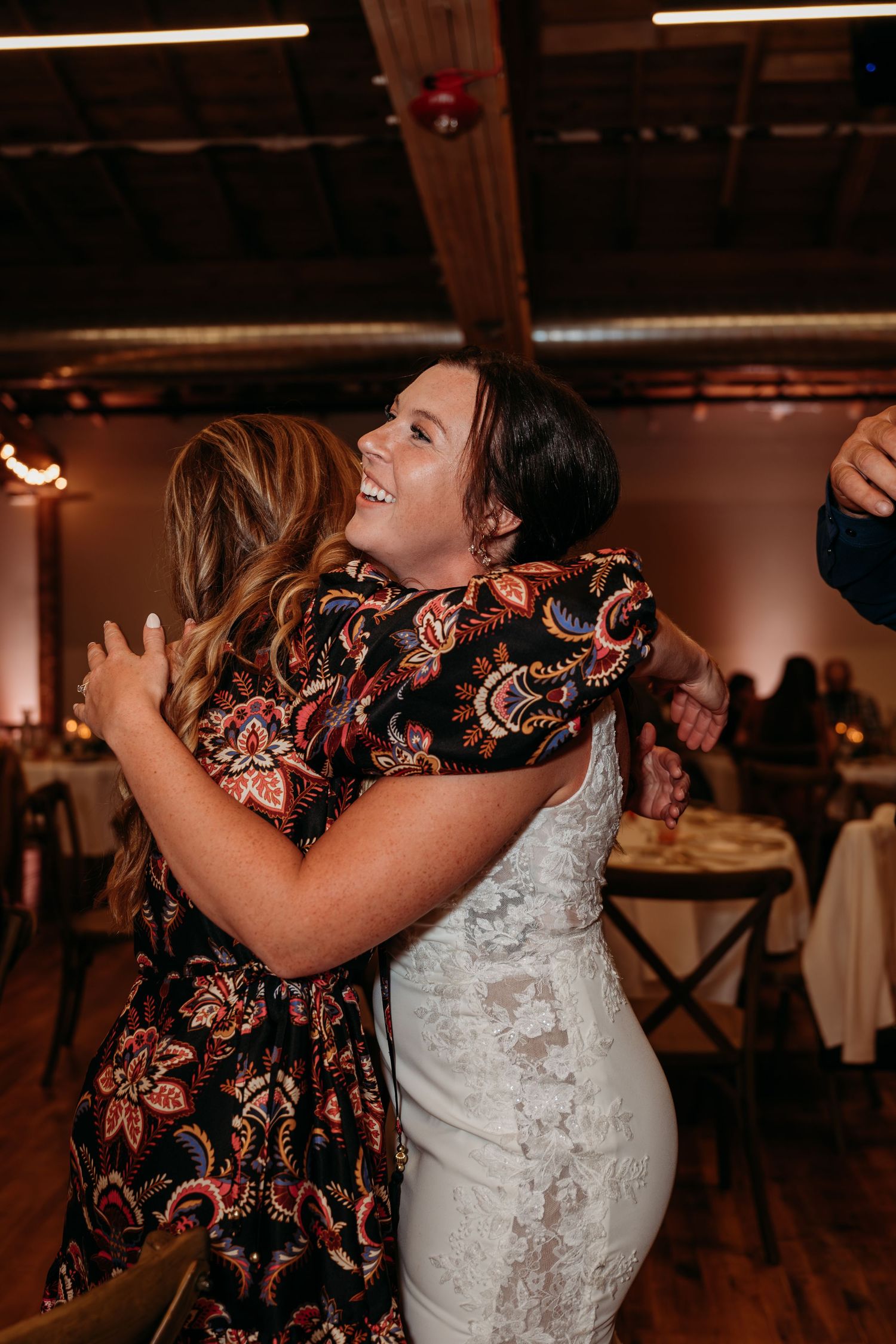 Bride in white lace dress hugging a guest in floral dress, laughing joyfully at a wedding reception.