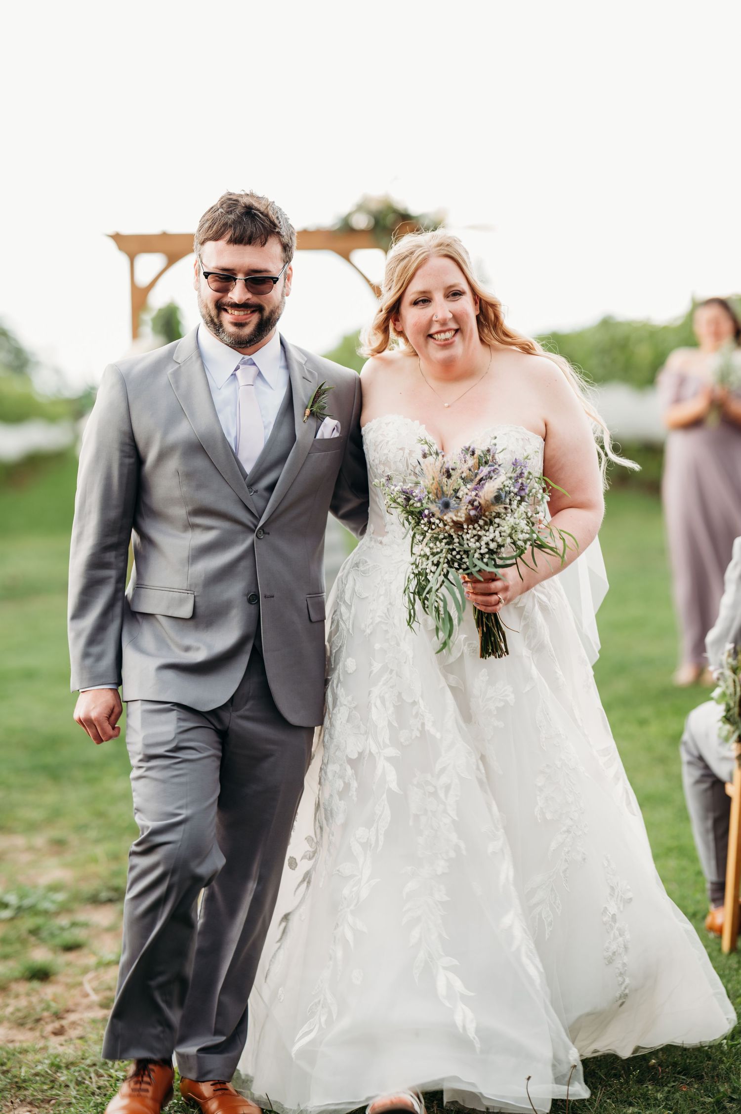 Smiling bride in white floral gown holding bouquet walks arm-in-arm with groom in gray suit after outdoor wedding ceremony.