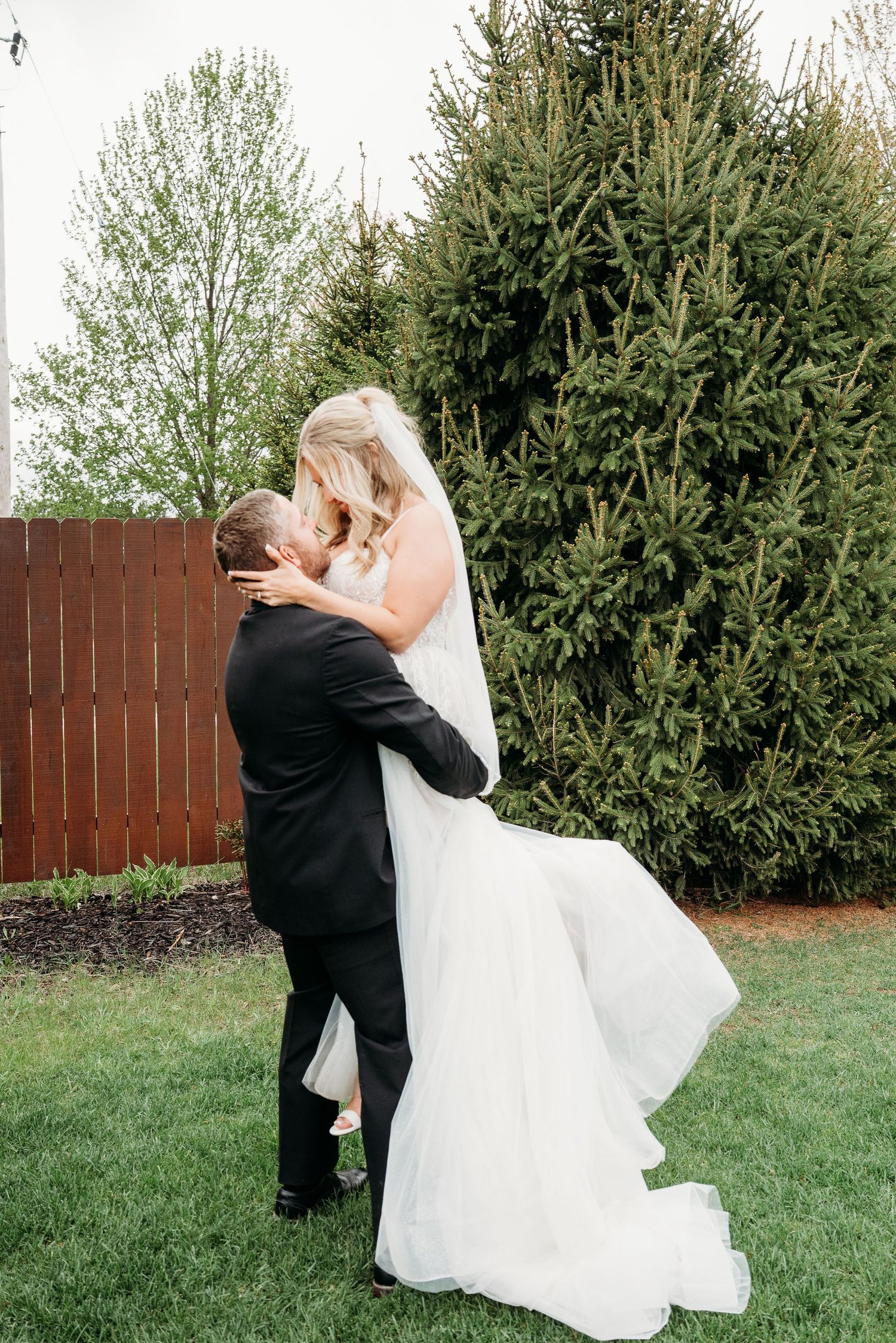 Groom lifting bride in white wedding gown outdoors near evergreen trees on lush green lawn.