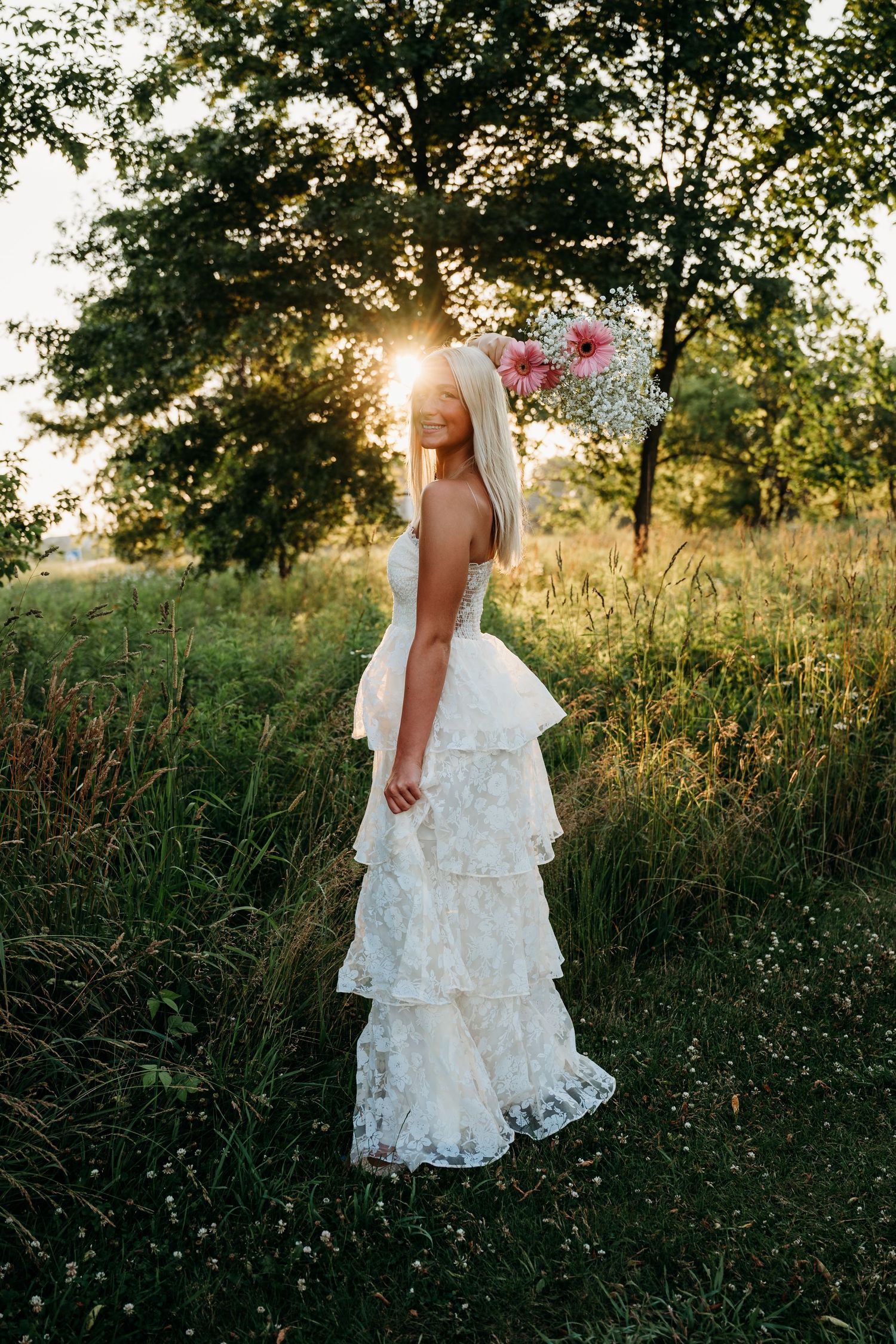 Bride in white lace tiered gown wearing floral crown stands in sunlit meadow at golden hour.