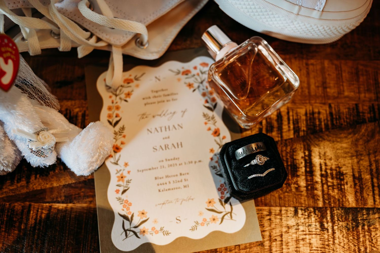 Wedding flat lay featuring Nathan and Sarah's invitation, gold perfume bottle, diamond ring, and wedding bands on a wooden surface.