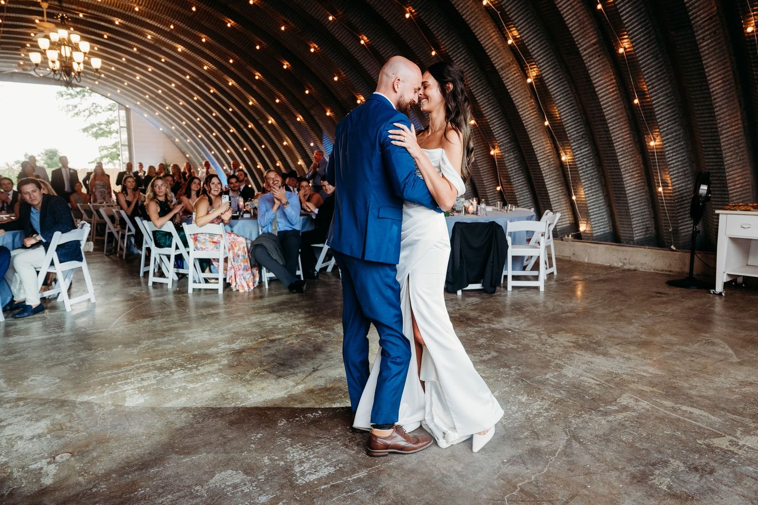 Bride and groom share first dance in rustic barn venue with string lights and seated wedding guests.