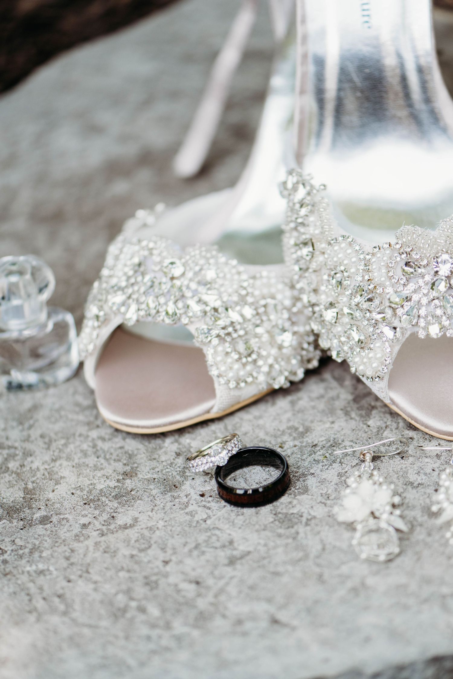 Wedding accessories featuring sparkly crystal bridal shoes and a black wedding band on stone surface.