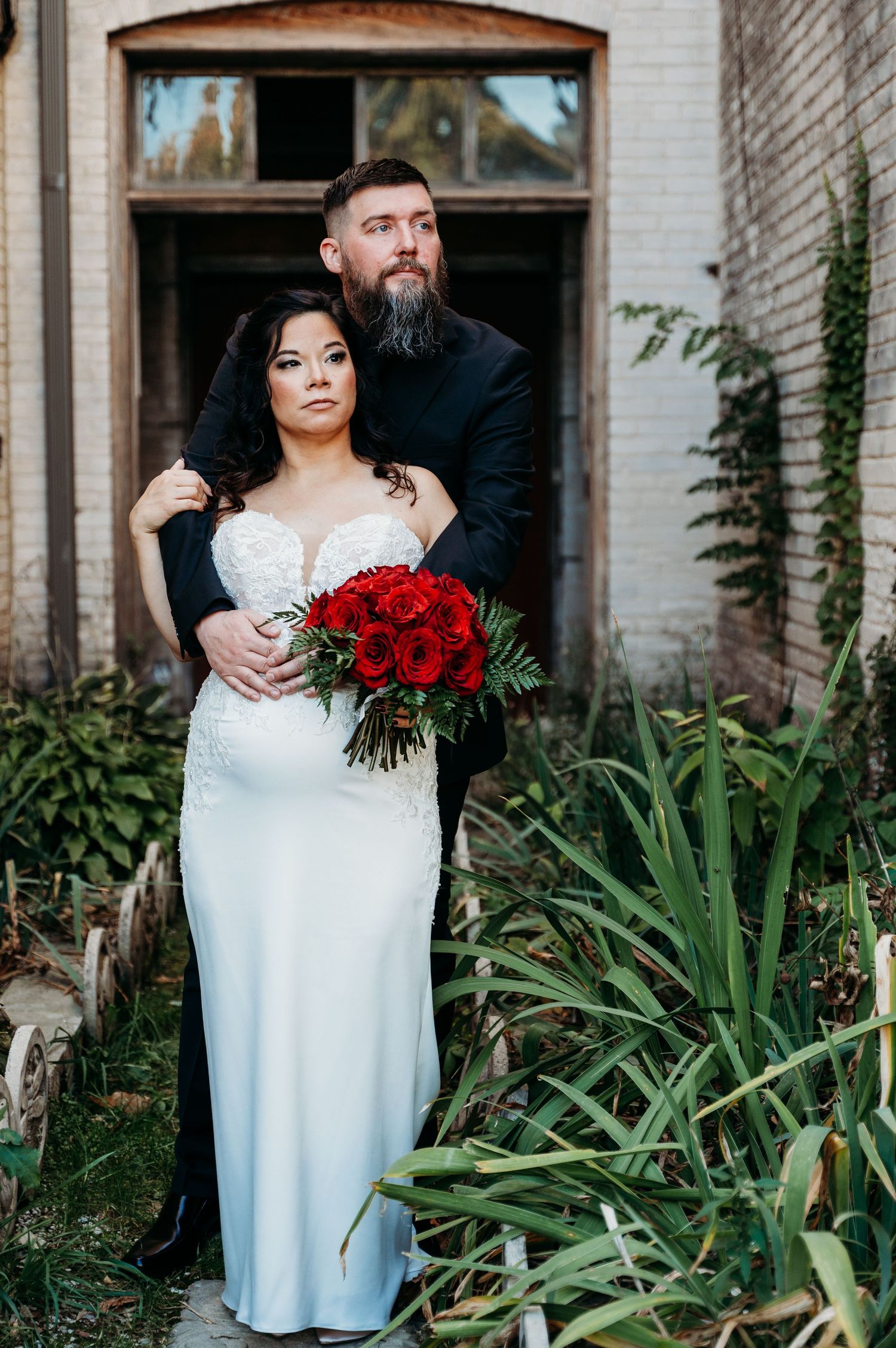 A couple in wedding attire embraces outside a rustic brick building with the bride holding a vibrant red rose bouquet.