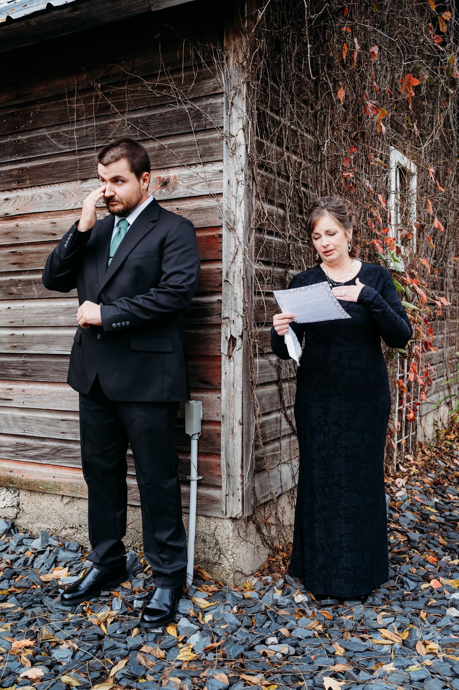 Two people in formal attire stand by a rustic wooden building reading papers on an autumn day.