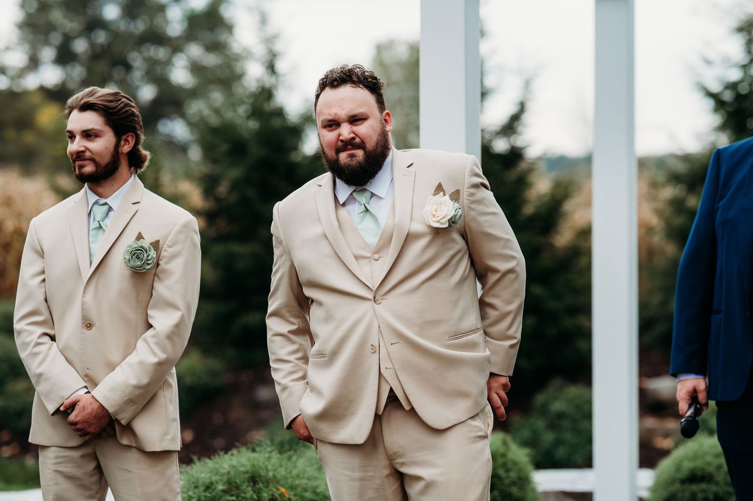 Groomsmen in tan suits and light blue ties stand together at an outdoor wedding ceremony.