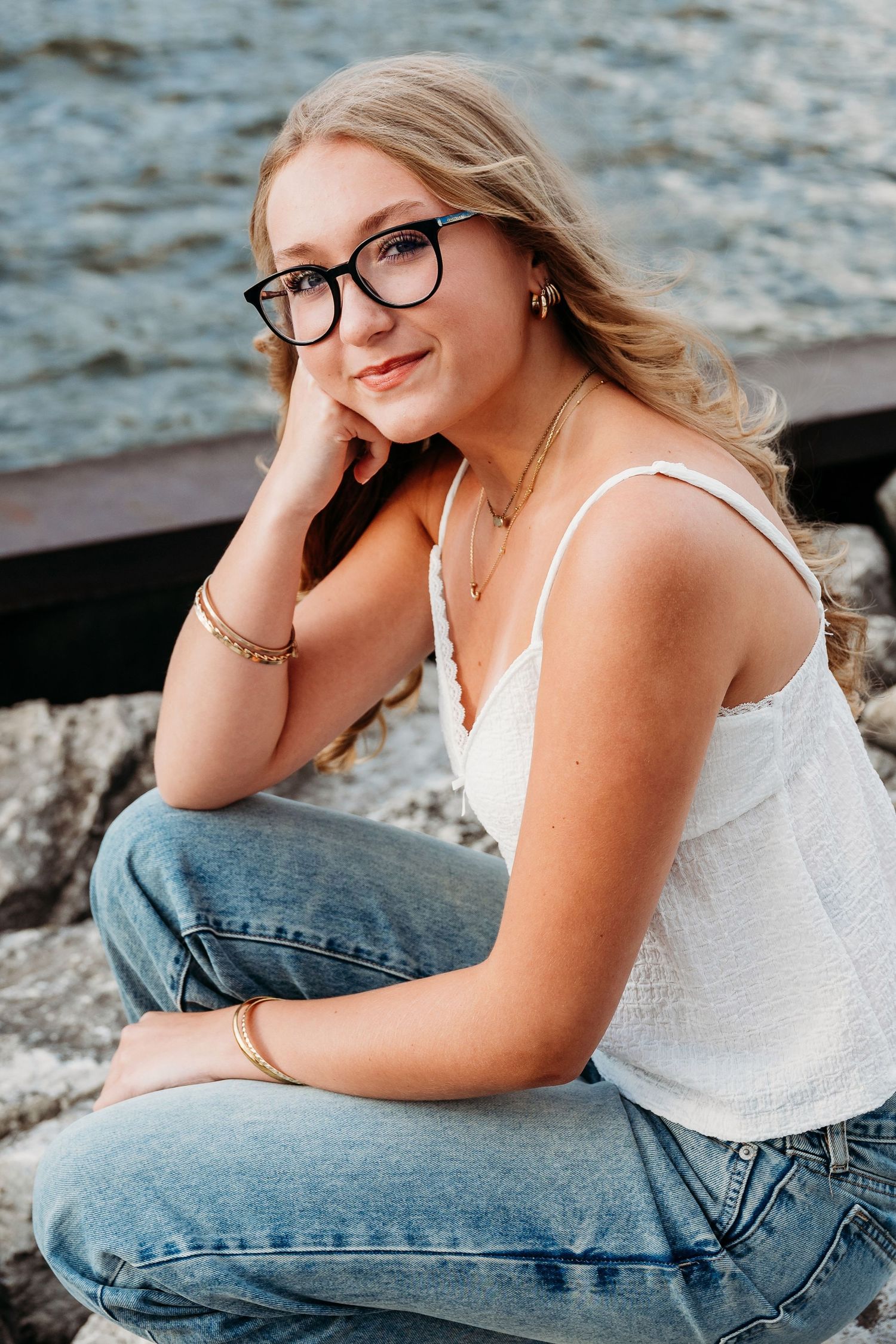 Casual waterfront portrait session featuring denim and white top while sitting on rocky shore.
