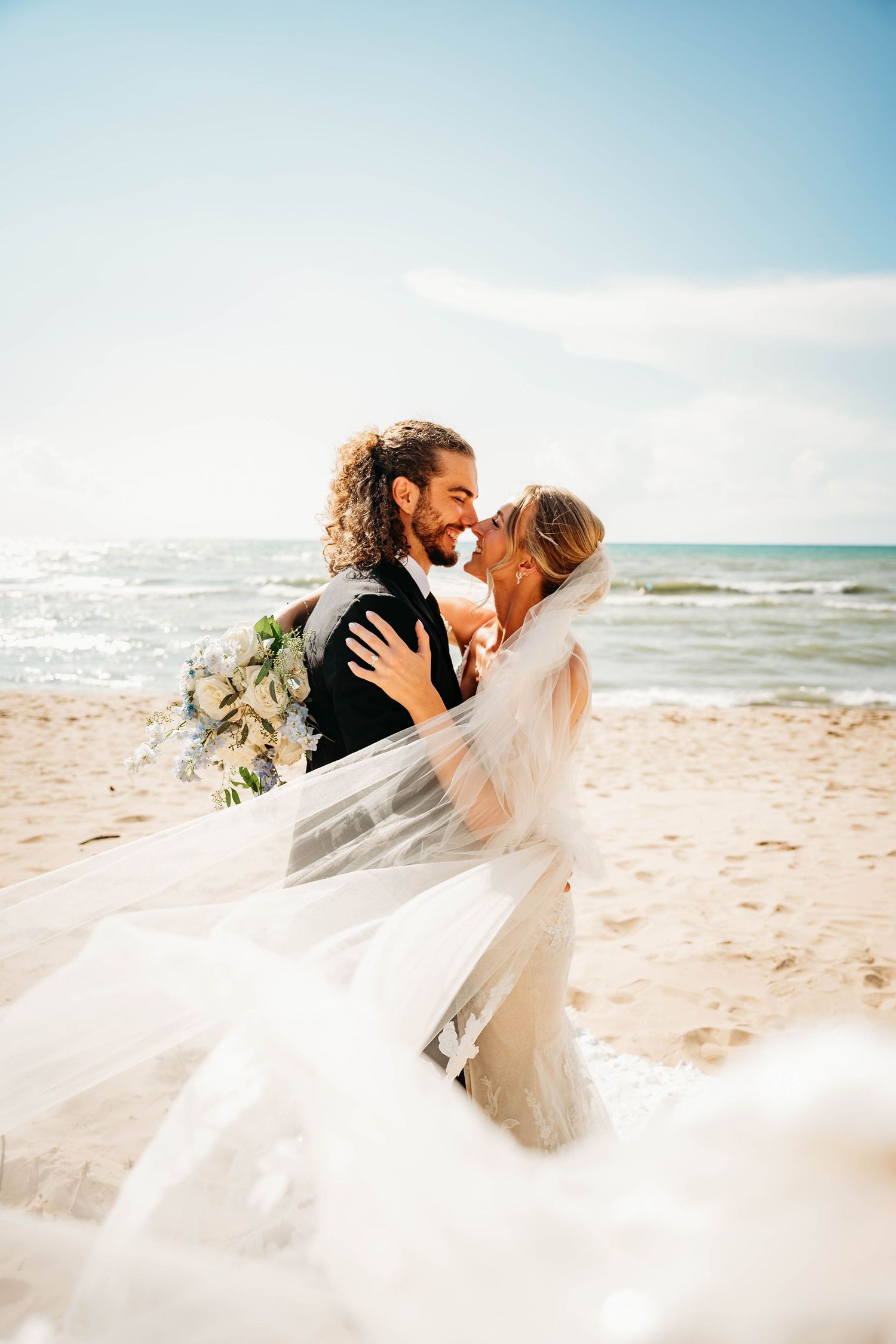 Bride and groom embrace on sandy beach, veil flowing in wind, ocean backdrop, sunny day wedding photo.