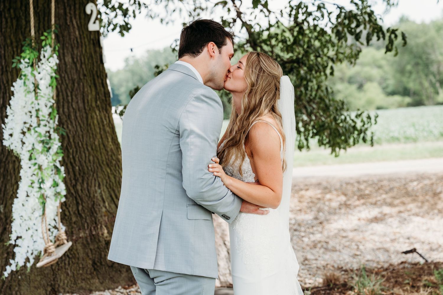 Bride and groom sharing a romantic kiss outdoors near a large tree with white floral decorations at their wedding.