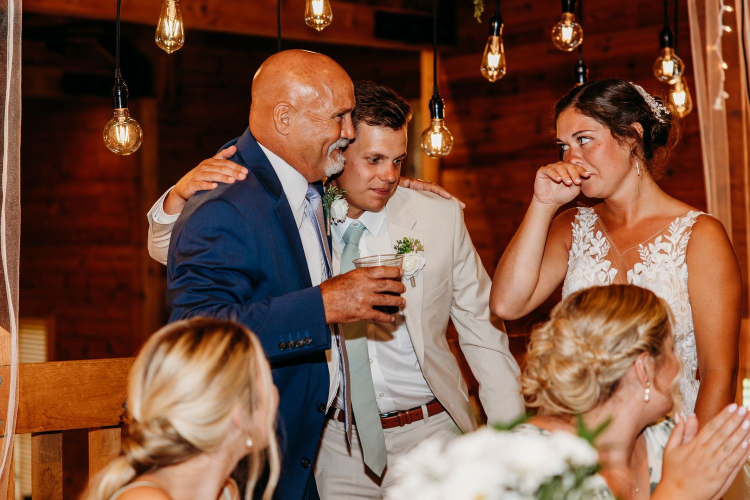 Emotional wedding reception moment with groom, bride, and older man sharing a heartfelt embrace in rustic barn venue.