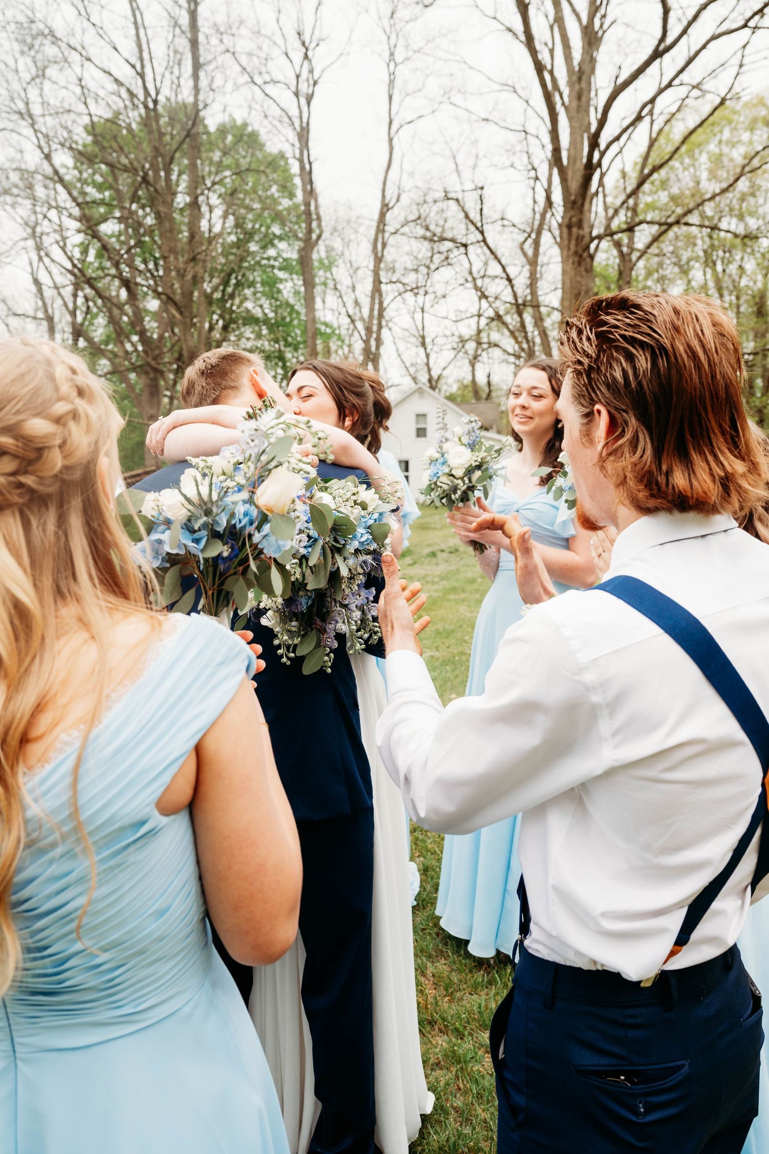 Wedding party in light blue dresses and navy suits sharing hugs outdoors, holding blue and white floral bouquets.