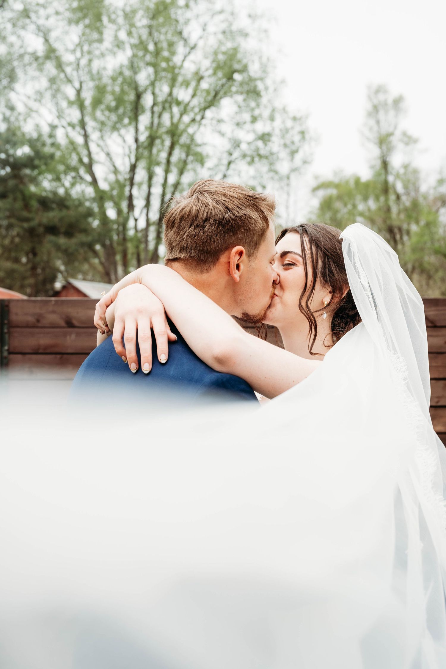 Bride and groom sharing a romantic kiss outdoors, with bride in white veil wrapping arms around groom in blue suit.