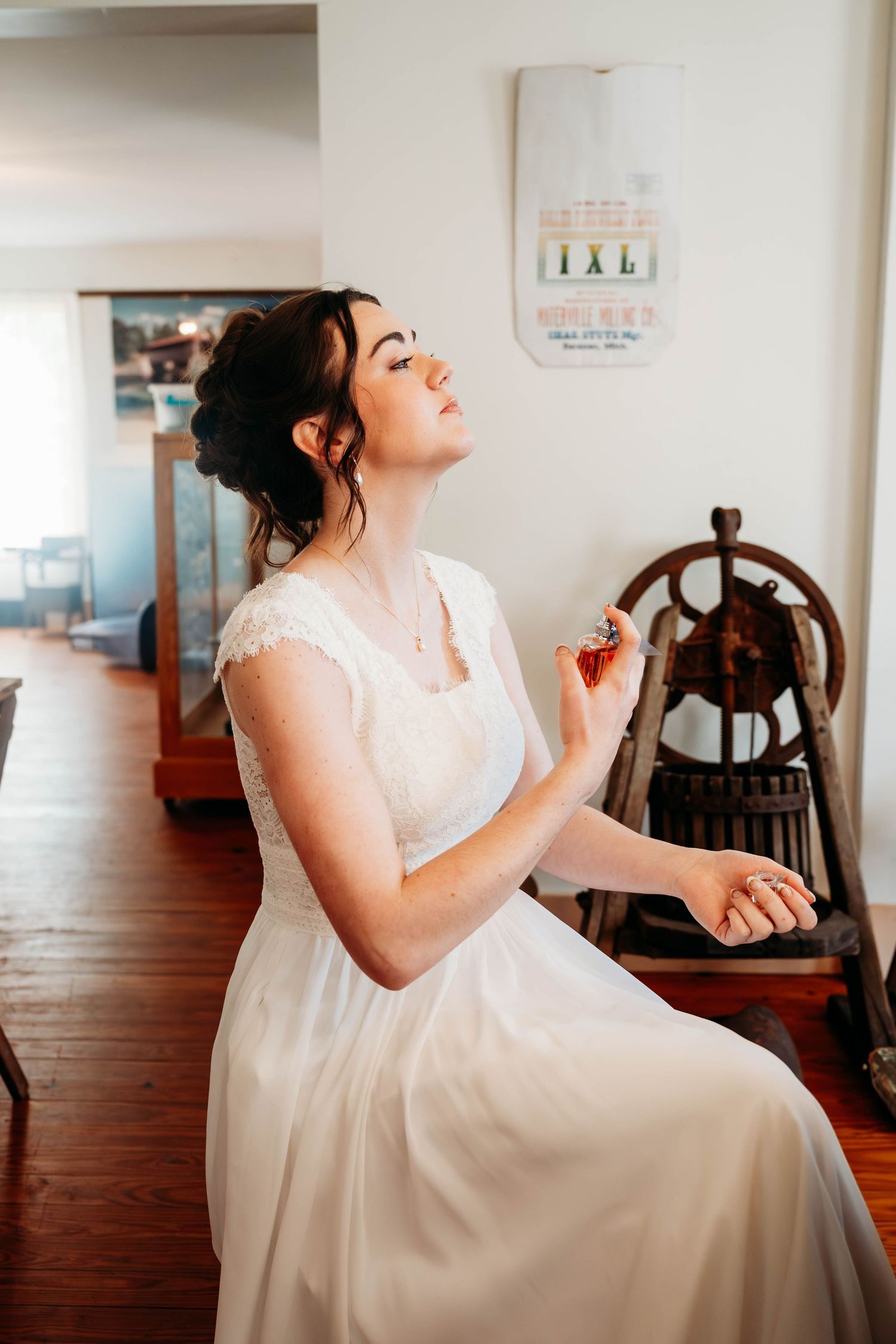 Bride in white lace gown applying perfume while seated, with eyes closed, in a rustic room with wooden decor.