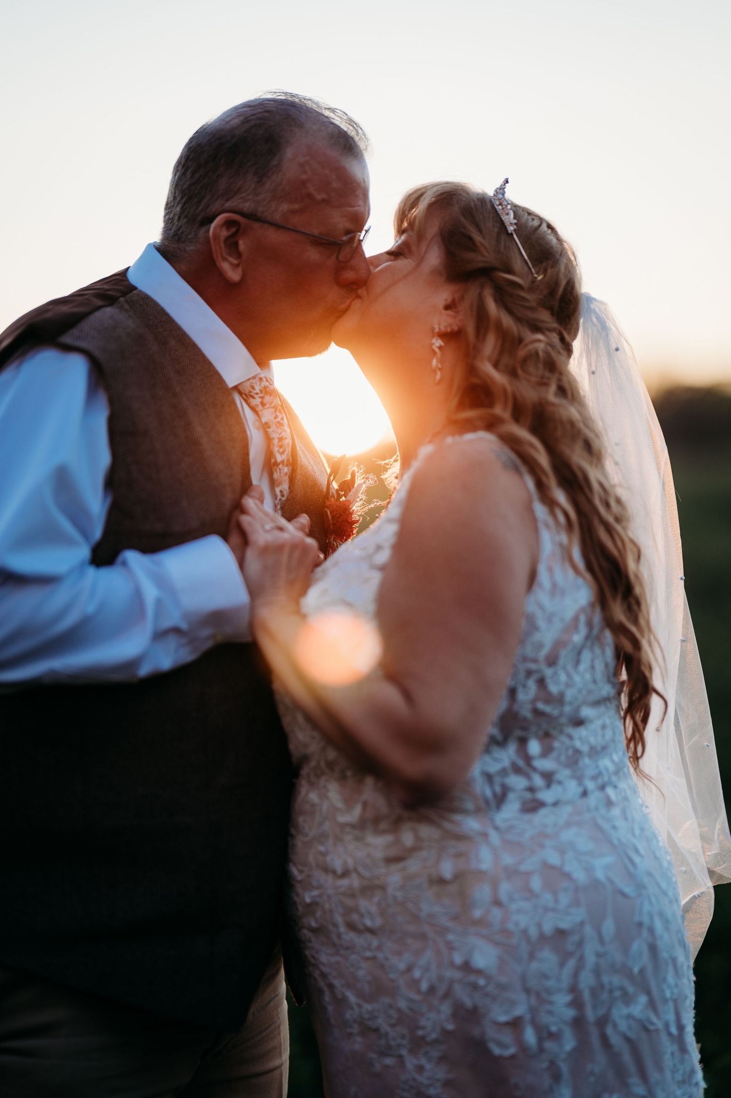 A couple in wedding attire share a romantic kiss at sunset with golden light glowing behind them.