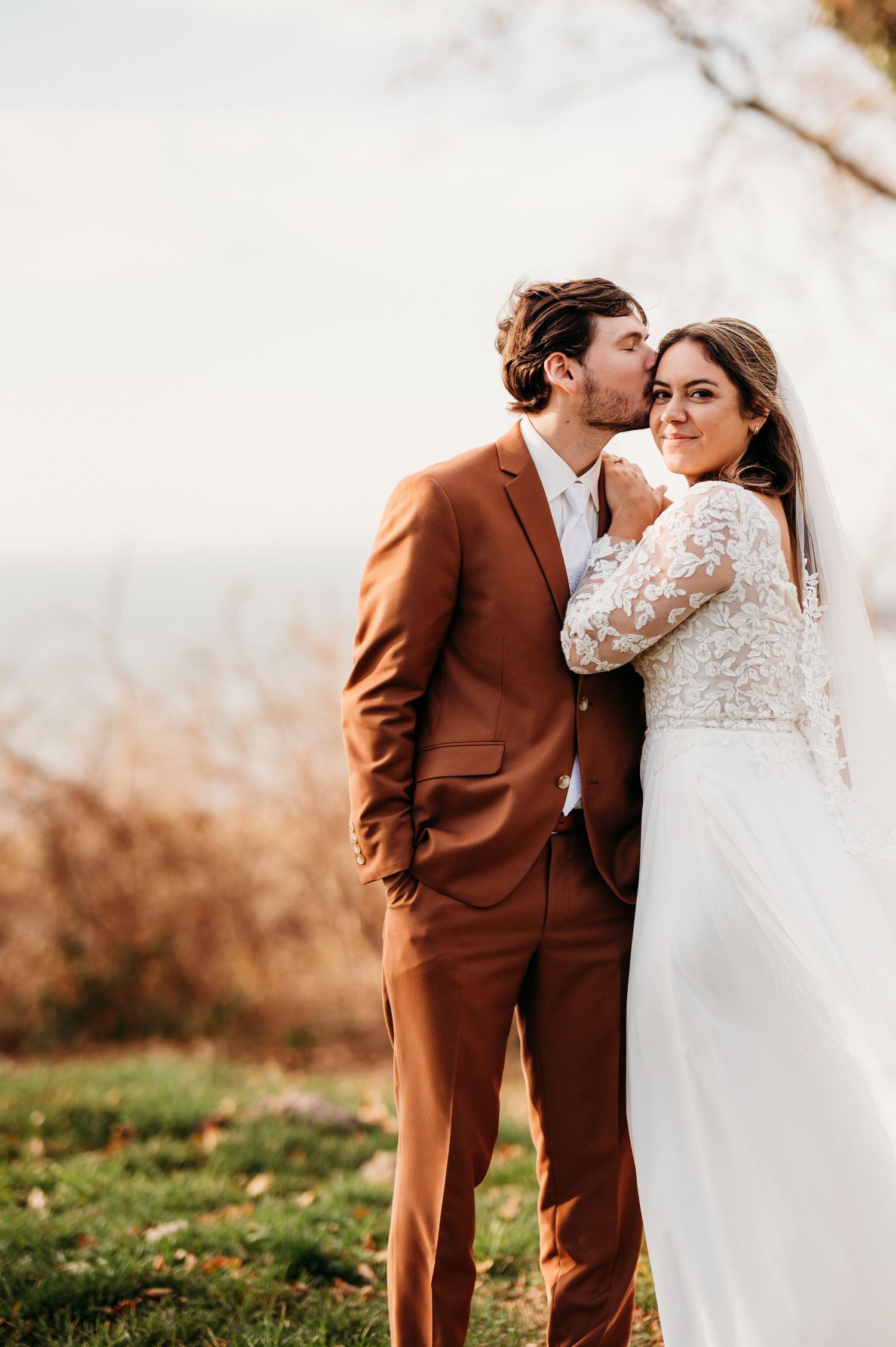A romantic wedding portrait showing a couple embracing outdoors, with the groom in a brown suit and bride in lace.