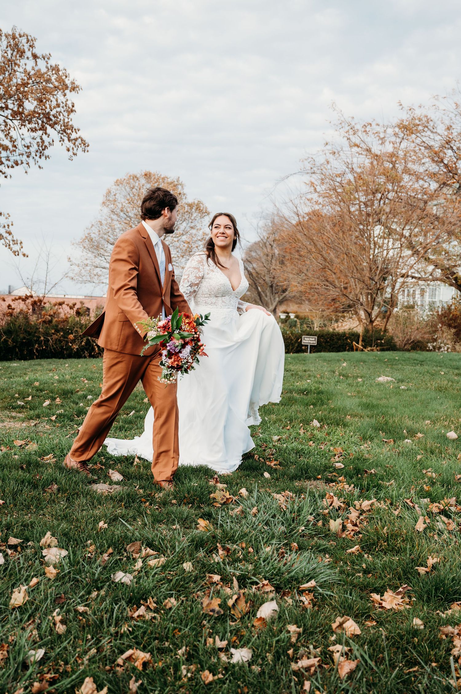 A couple in autumn wedding attire playfully runs through a grassy field covered in fallen leaves.