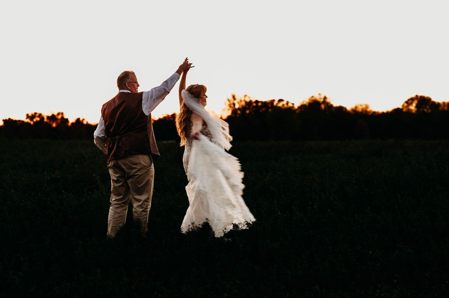 A couple holds hands with raised arms against a sunset sky, the bride wearing a flowing white wedding dress.
