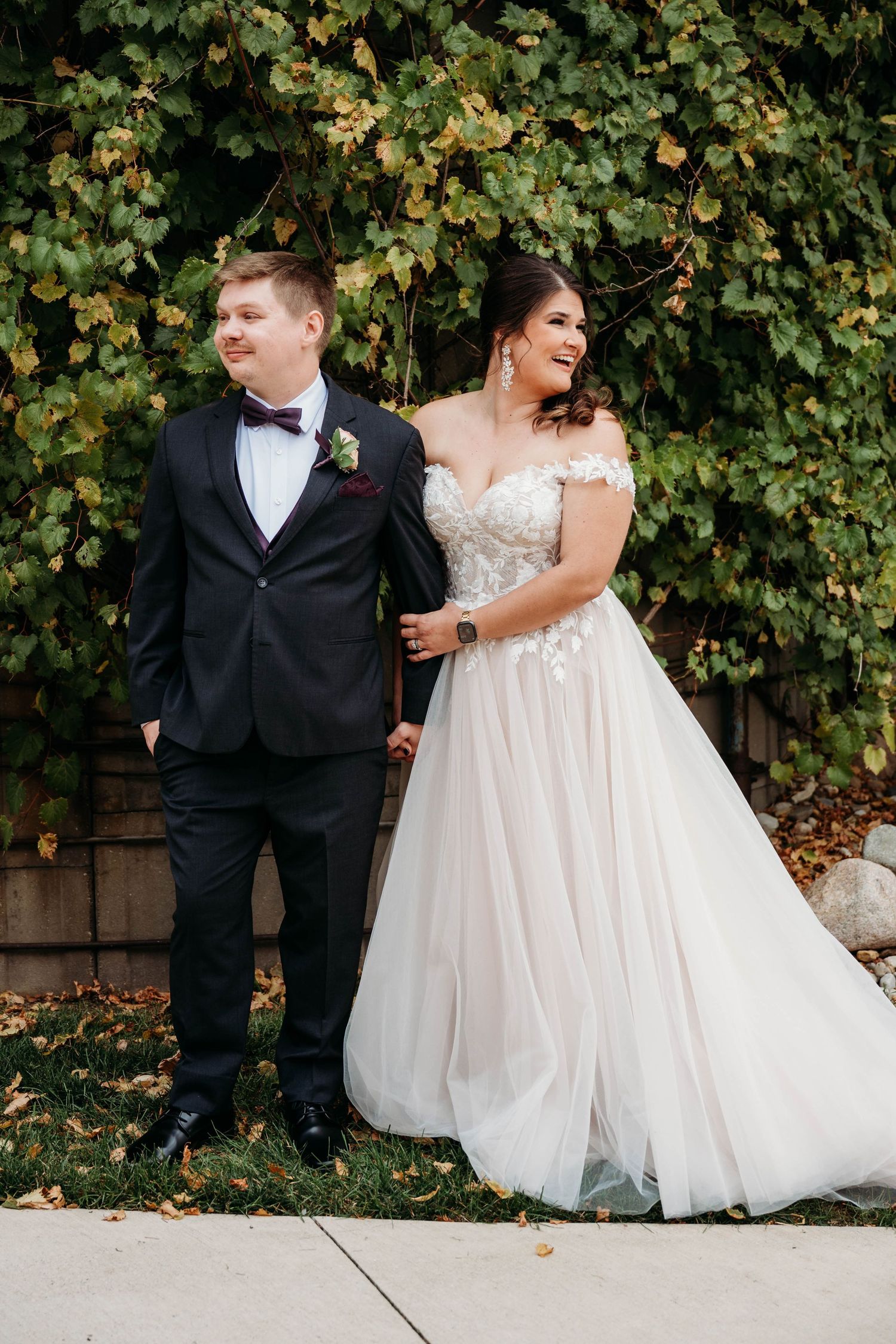 A joyful wedding couple shares a candid moment together against a backdrop of lush green vines and autumn leaves.
