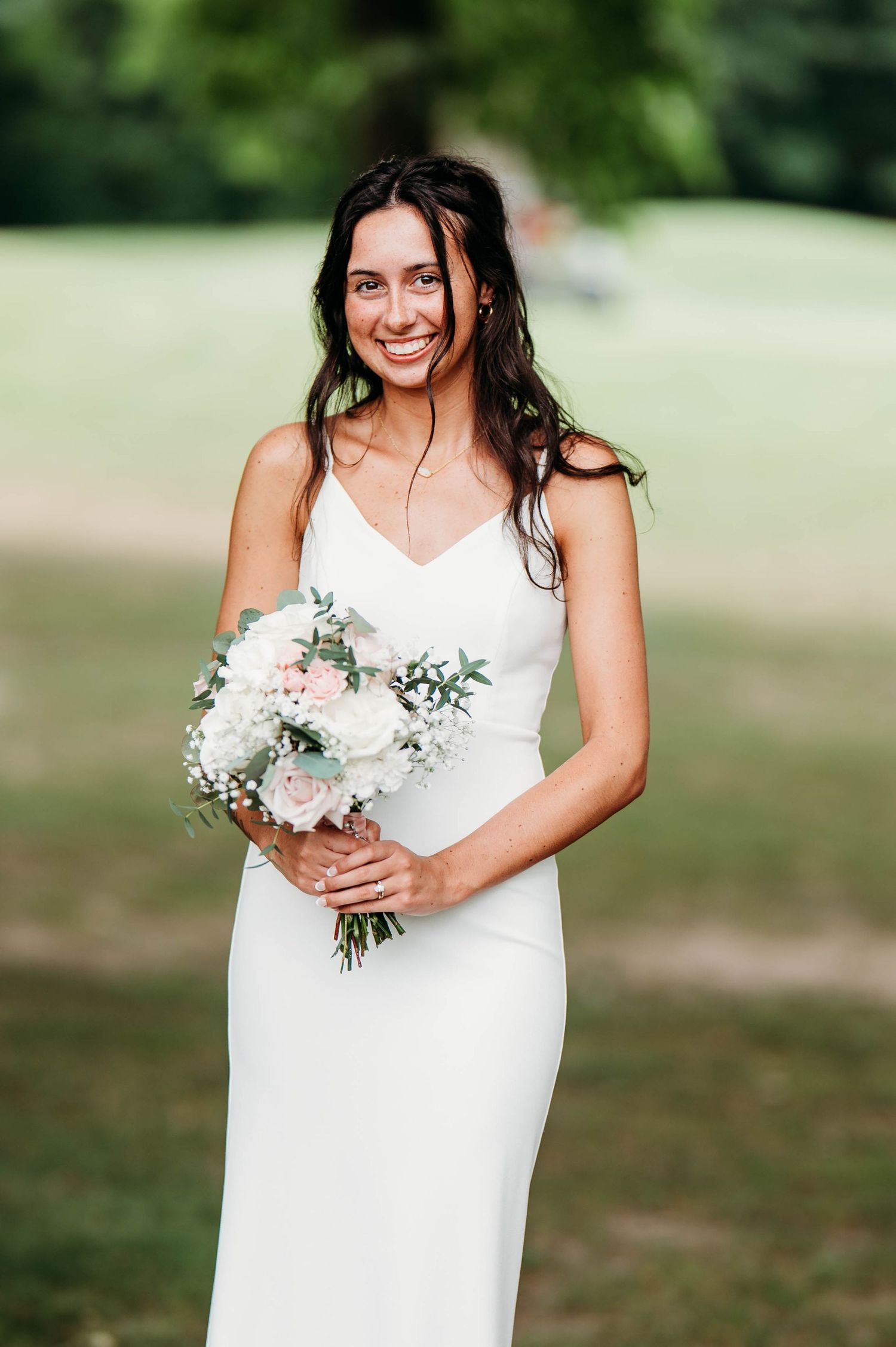 A bridal bouquet with white roses and greenery is held against a white dress outdoors.