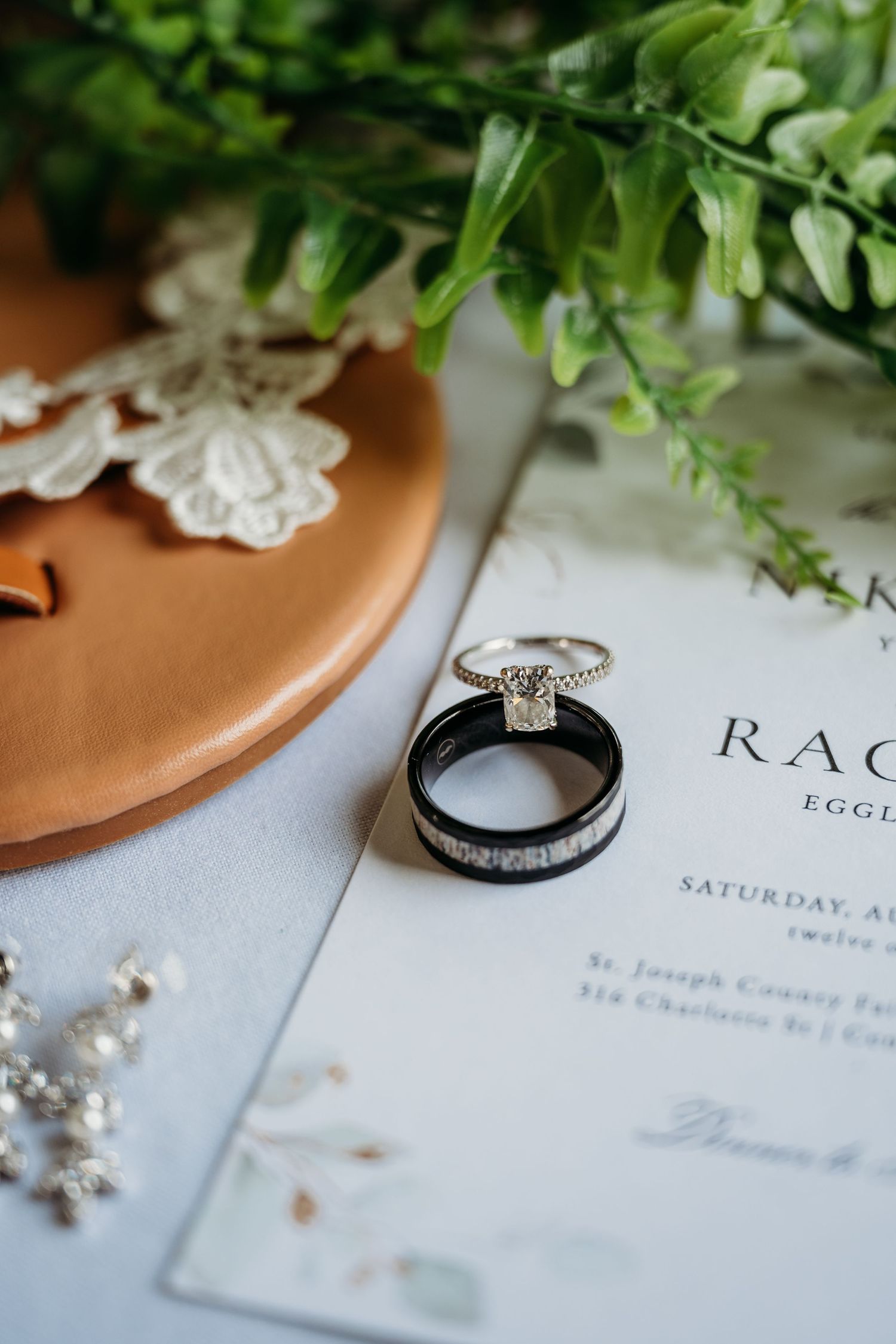Wedding rings and invitation details arranged with greenery and soft lighting on a light blue surface.