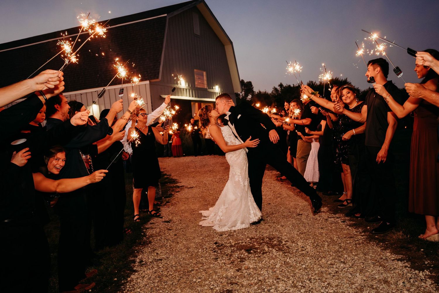 Wedding couple shares a romantic kiss under a sparkler tunnel created by guests at a rustic barn venue at night.