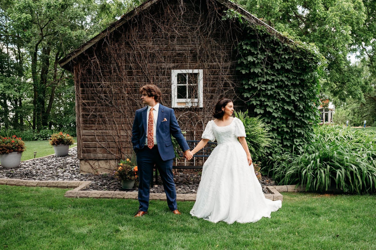 Bride and groom holding hands walking on green lawn beside rustic ivy-covered cottage surrounded by lush trees.