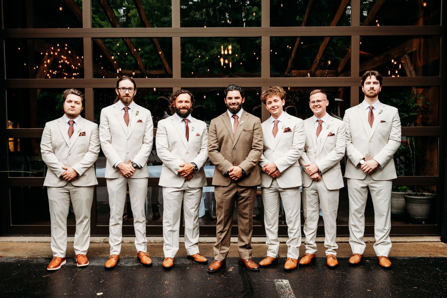 Groom in tan suit stands with six groomsmen in matching light gray suits and red ties outside a modern venue.