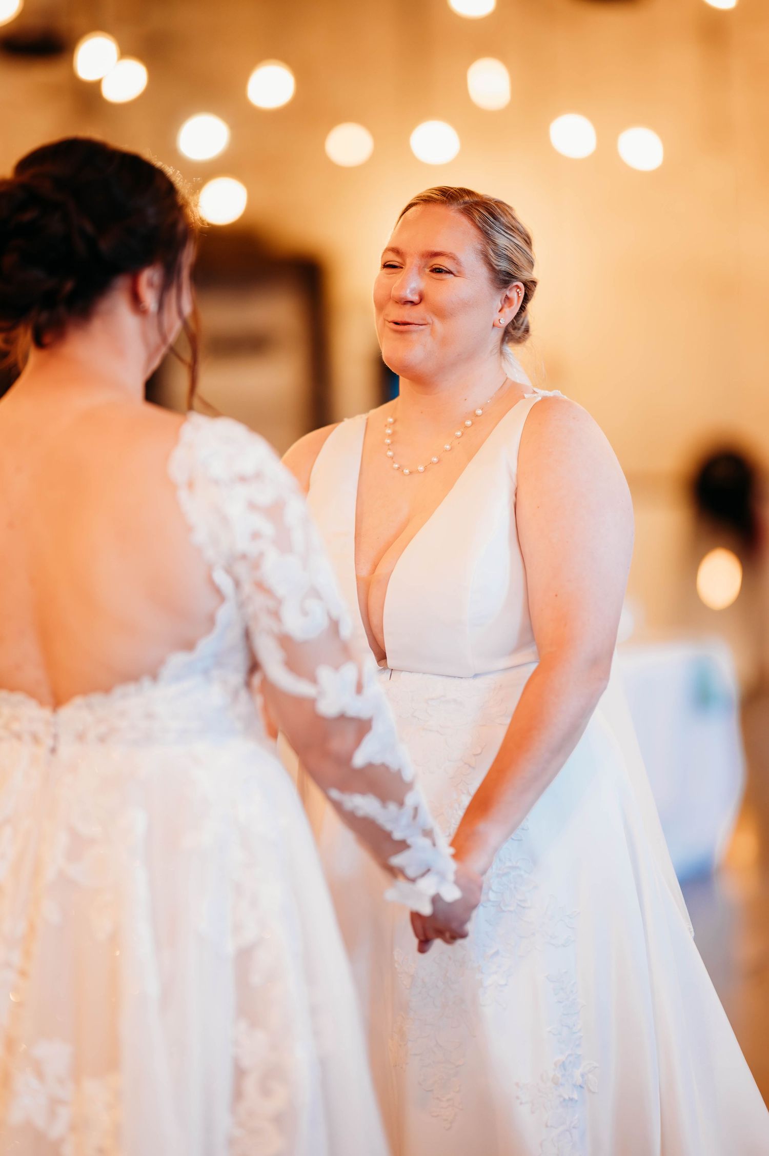Bridesmaids in elegant white wedding dresses dancing and celebrating at a reception with warm lighting.
