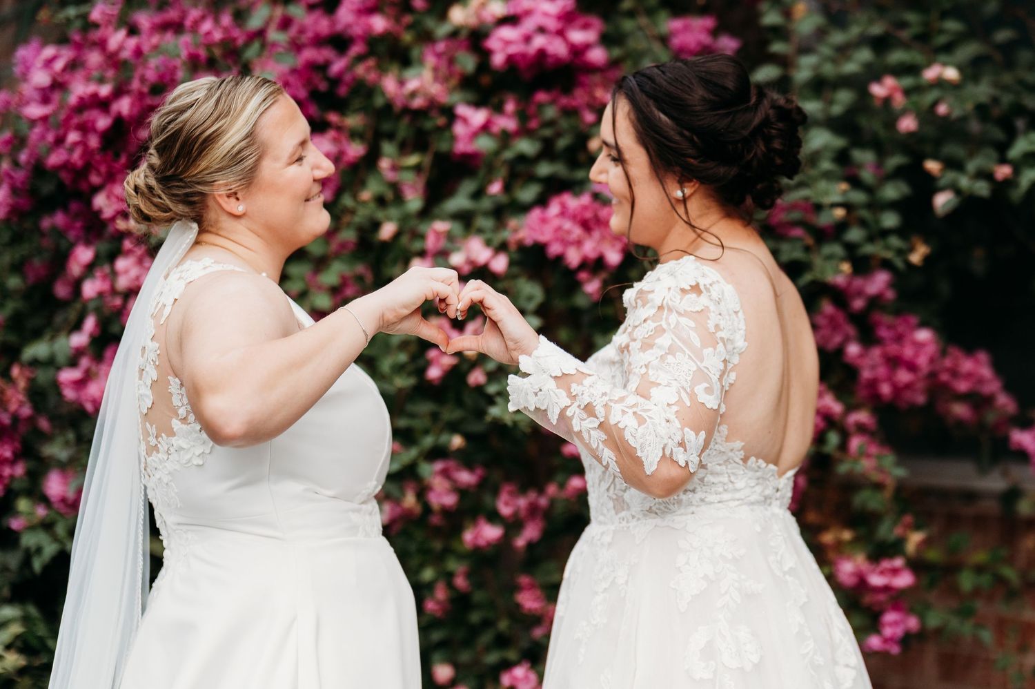 Two brides share a joyful dance moment together in front of vibrant pink flowers during their wedding celebration.