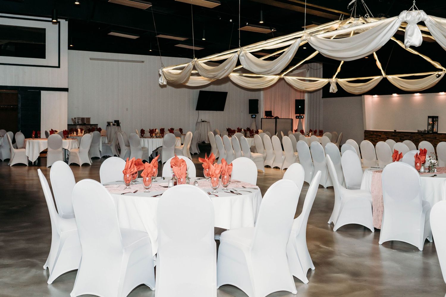 Wedding reception venue decorated with white chair covers and coral accents under draped fabric ceiling details.