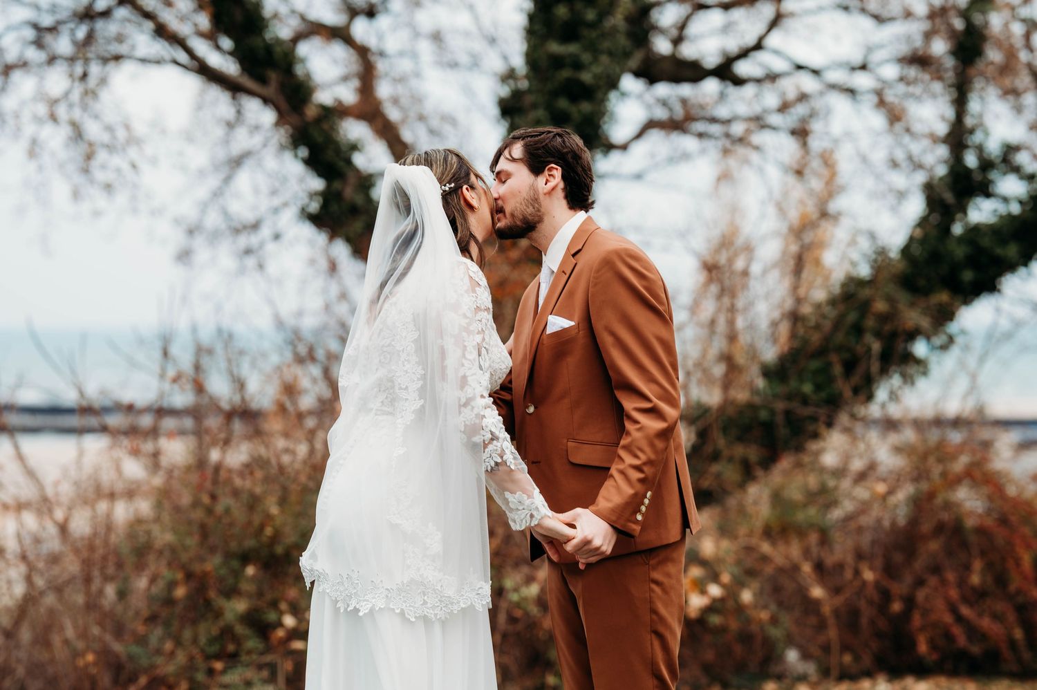 A couple in wedding attire shares a tender moment outdoors during an autumn day with bare trees in the background.