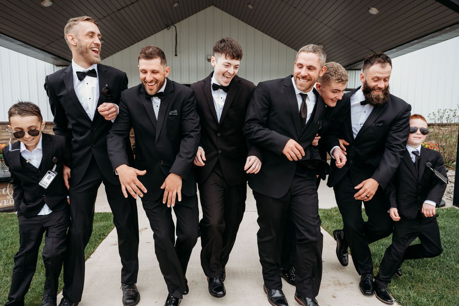 A group of groomsmen in black tuxedos perform a synchronized jumping pose outdoors.