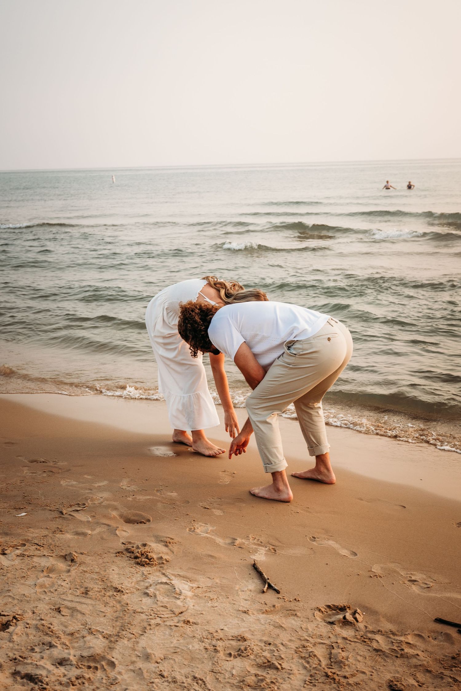 Couple in matching white outfits playfully bending down to write in the sand at the beach.