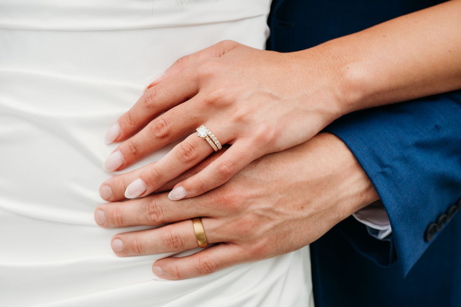 Two hands wearing wedding rings rest gently against a white and navy backdrop, showcasing their matching bands.