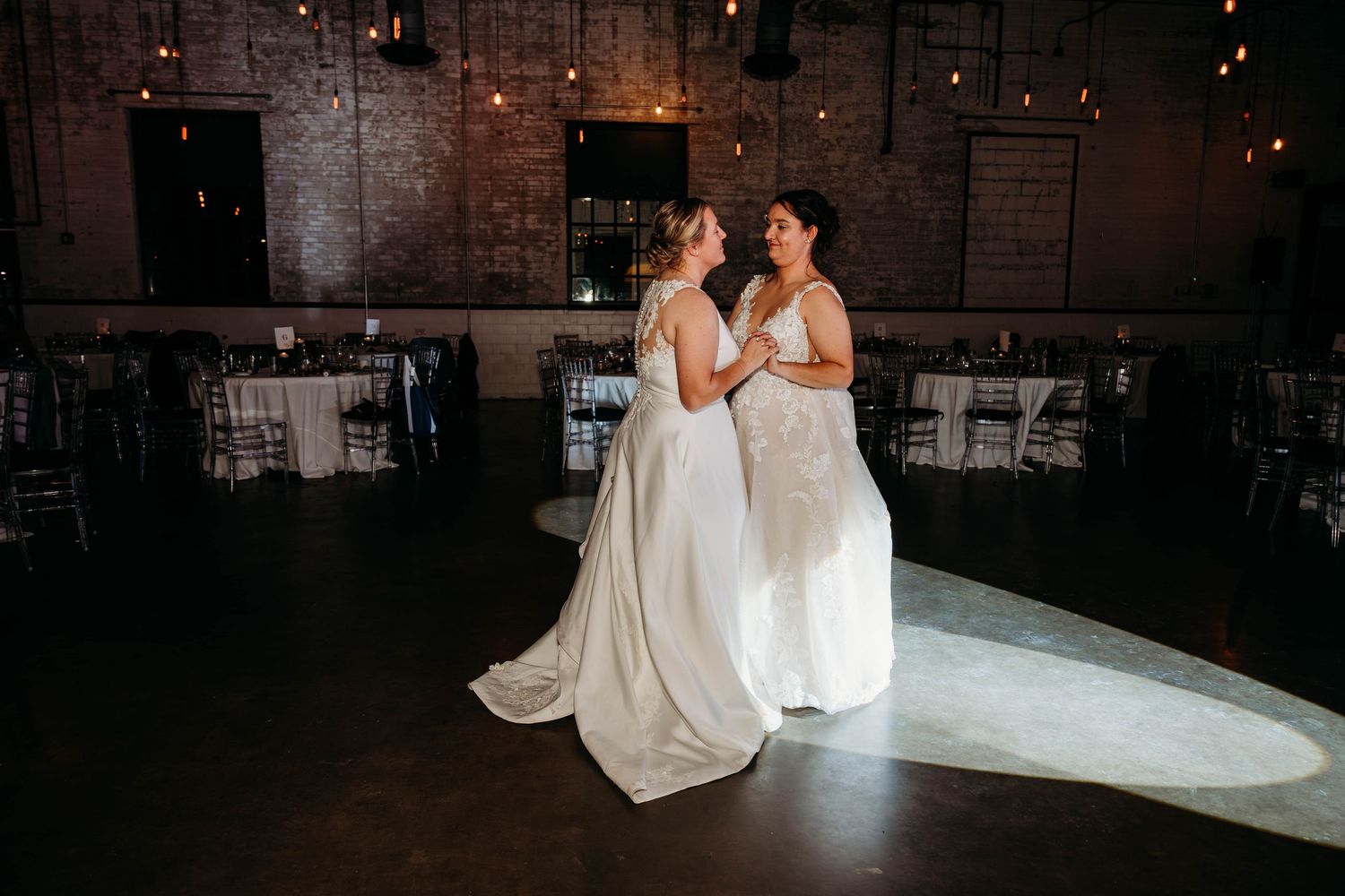 Two brides in white wedding gowns share a first dance in a romantic industrial venue with string lights and brick walls.