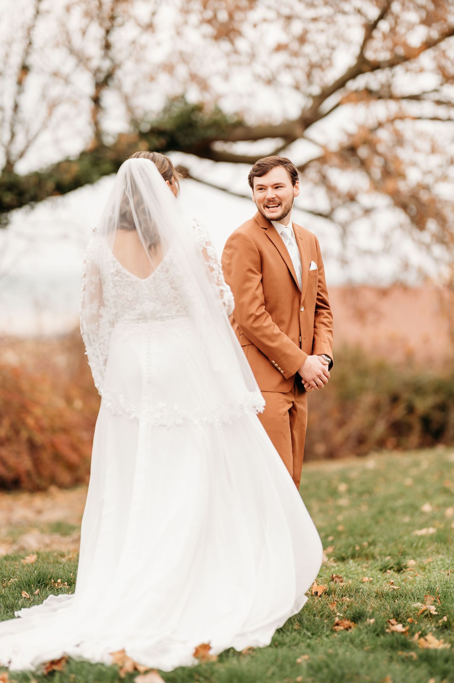 Groom in rust brown suit smiling at bride in white lace wedding dress and veil during fall outdoor first look.