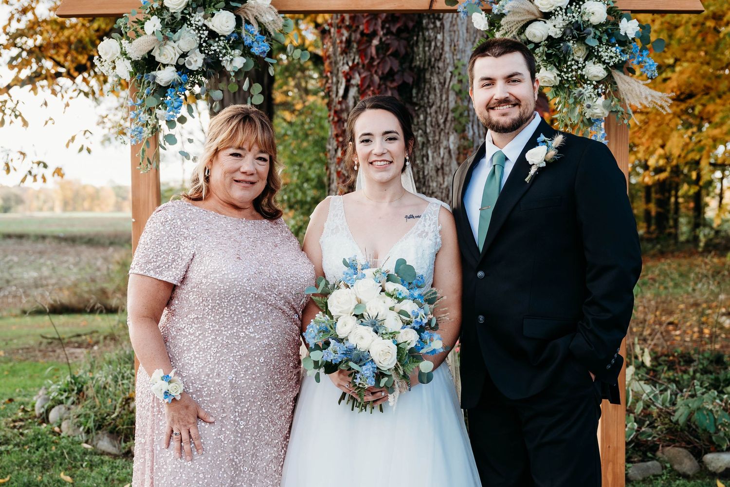Bride and groom pose with family member at outdoor wedding ceremony, bride holding blue and white floral bouquet.