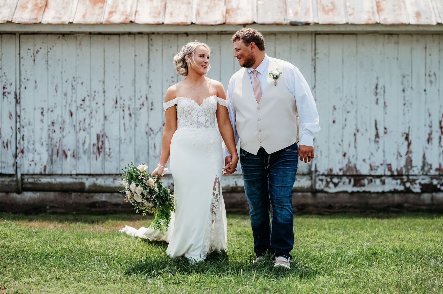 Bride and groom holding hands walking in front of a rustic barn, bride wearing lace off-shoulder gown holding bouquet.