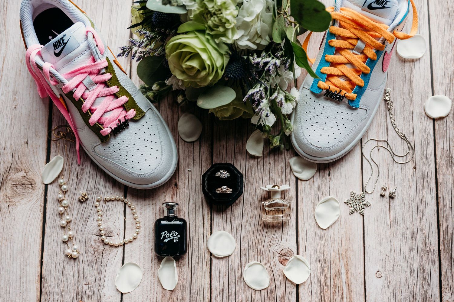 Nike sneakers with colorful laces beside a floral bouquet, rings, jewelry, and perfume on a wooden surface.
