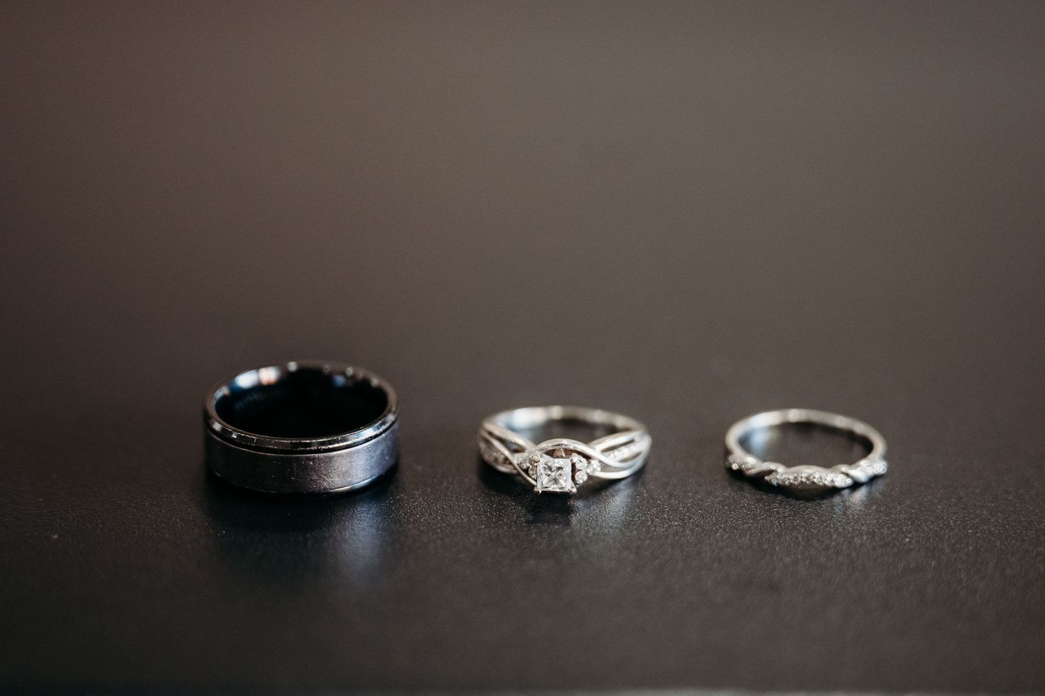 Three silver wedding rings displayed on dark surface, including mens band, diamond engagement ring, and eternity band.