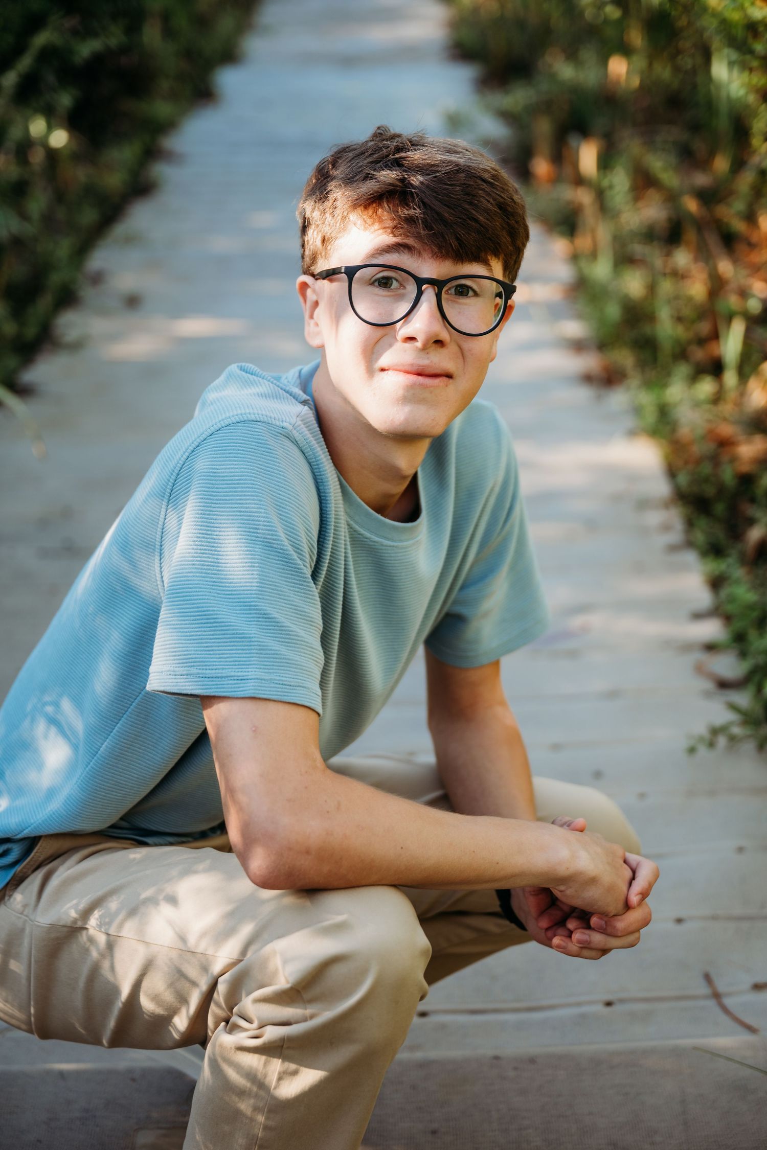 Person wearing glasses and blue t-shirt sitting outdoors on sidewalk smiling with casual pose.