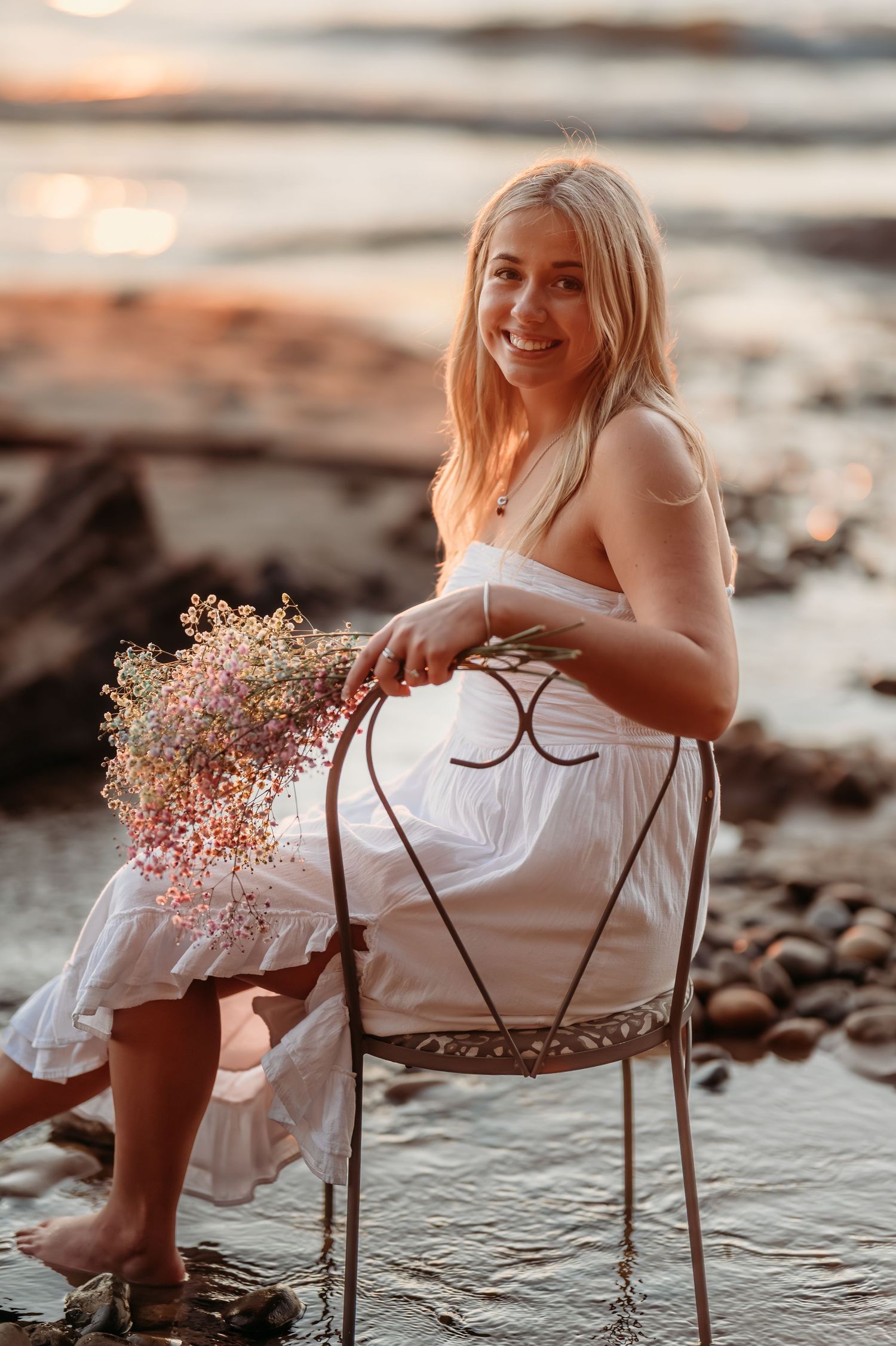 Woman in white dress and dusty rose bouquet sitting in vintage metal chair during sunset beach photoshoot.
