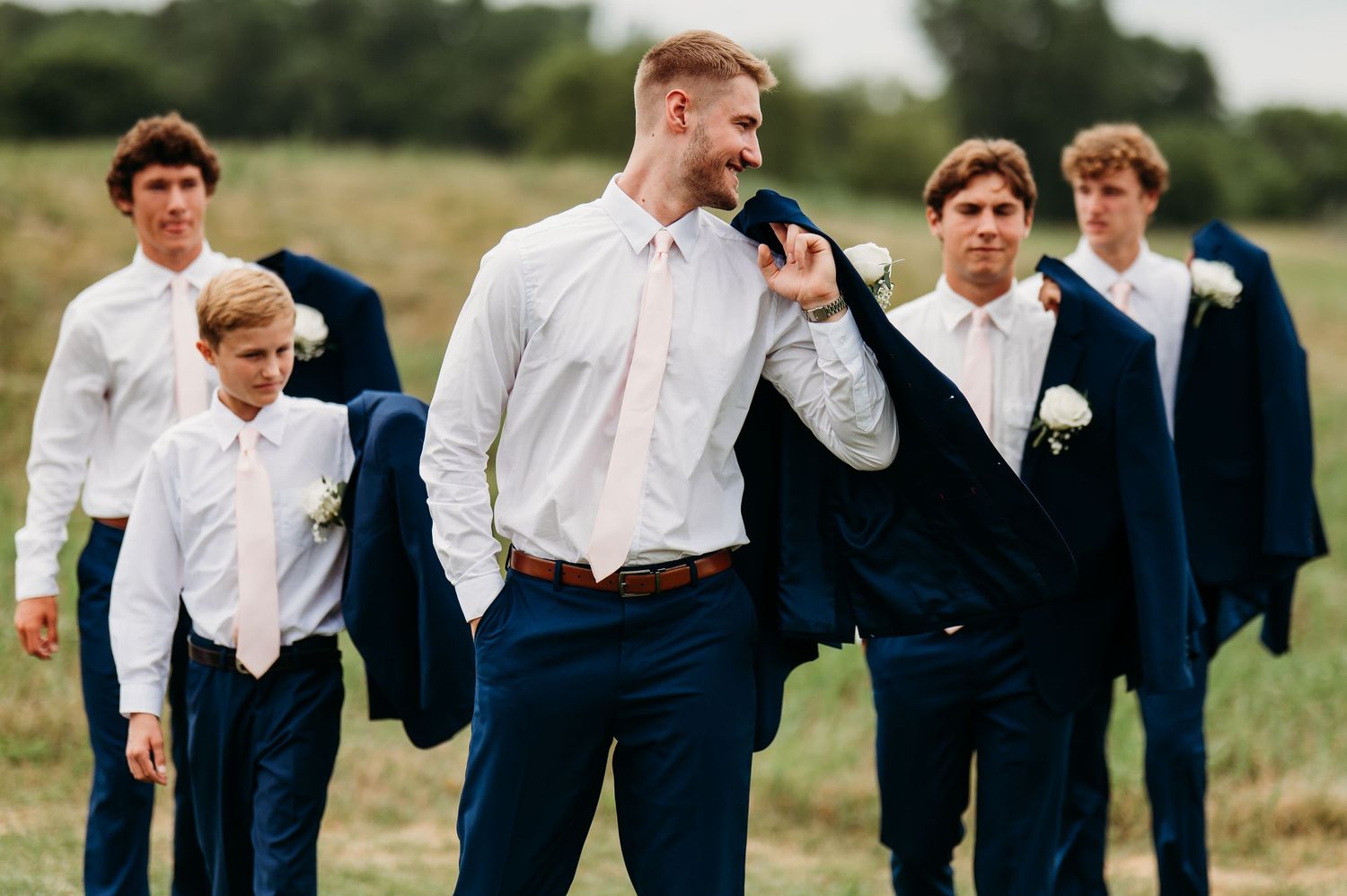 Groomsmen in navy suits and white shirts share a candid laughing moment together in an outdoor field setting.