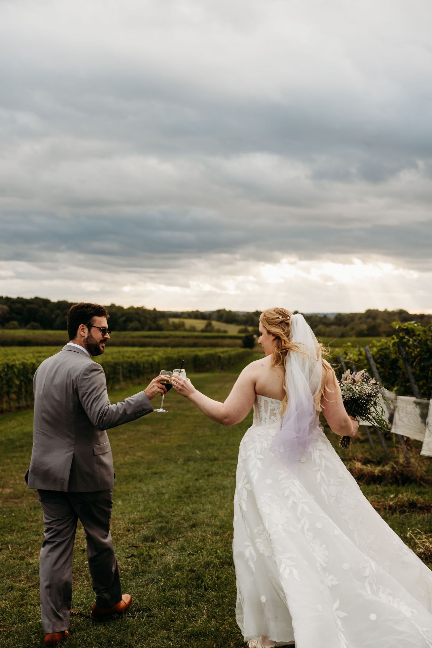A couple in wedding attire toasts with champagne glasses in a scenic outdoor field under cloudy skies.