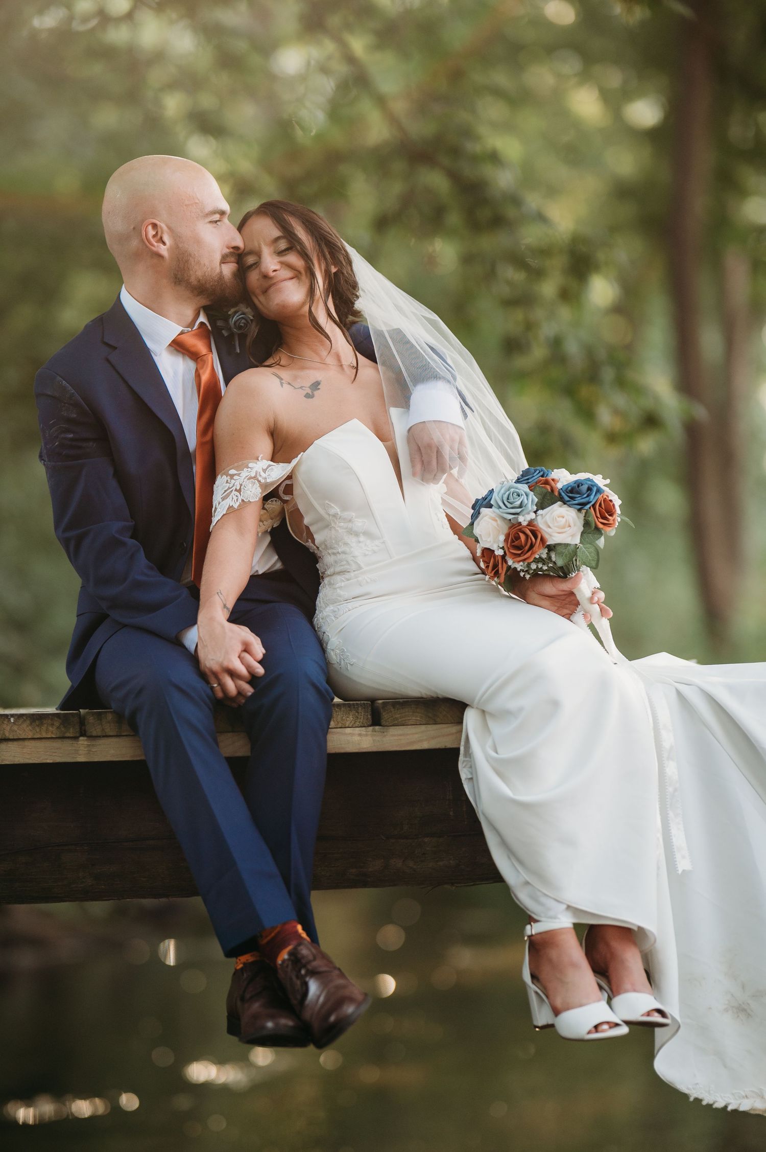 A romantic wedding couple sits together on a wooden bench in a garden setting wearing navy suit and white dress.
