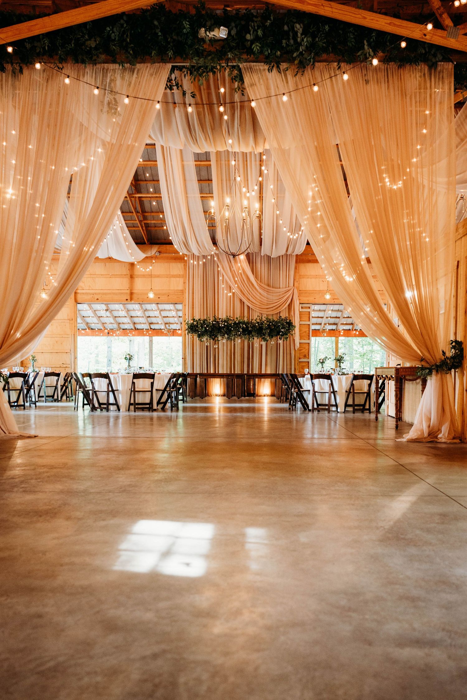 Elegant wedding venue interior with sheer ivory drapes, string lights, and wooden beams in a rustic barn setting.